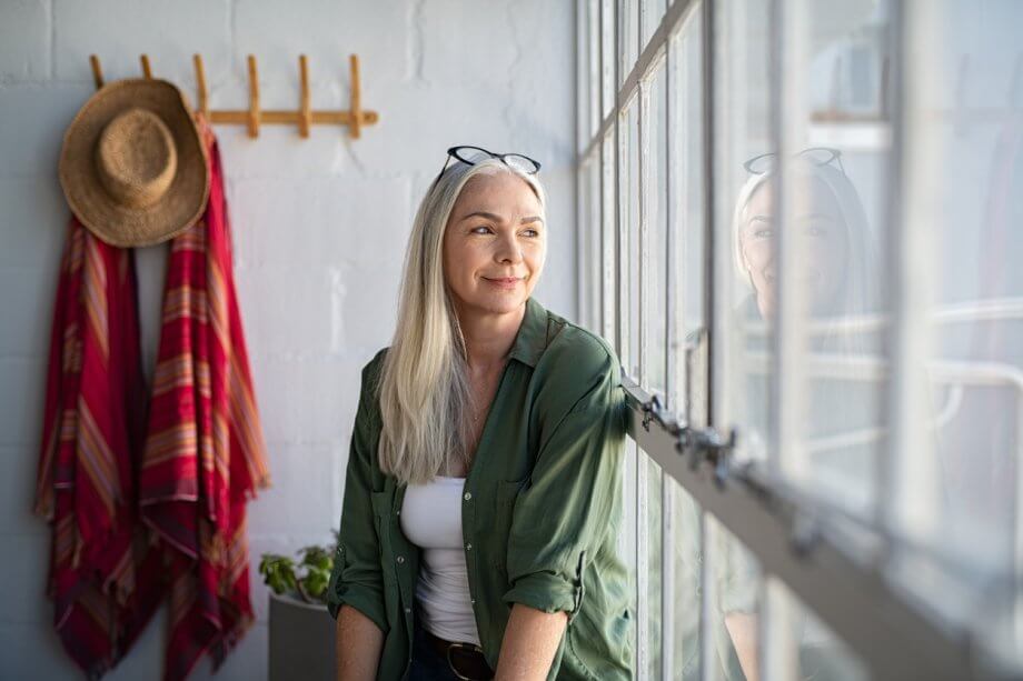 Older woman with long white hair looking pensively out of the window of her home